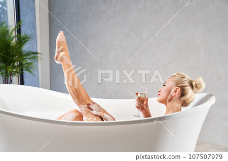 Relaxed blonde woman with hair bun, taking warm bath at home in evening. Side view of female lying in bathtub, with leg sticking out of bubble bath, in front of grey wall. Concept of spa and relax. 107507979