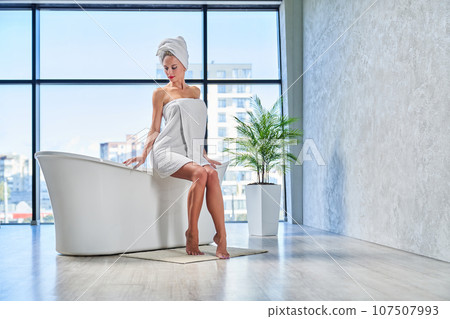 Young lady with hair wrapped in white towel looking down, while sitting on bathtub edge. Low angle view of pretty woman resting after taking bath in morning, in front of window at home. Daily routine. Young lady with hair wrapped in white towel looking down, while sitting on bathtub edge. Low angle view of pretty woman resting after taking bath in morning, in front of window at home. Daily routine. 107507993