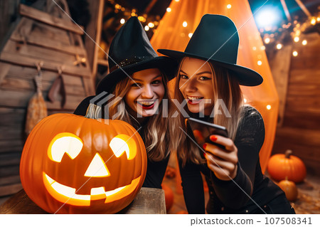 Two young women friends wearing witch hats having fun and posing with a jack-o'-lantern at Halloween 107508341