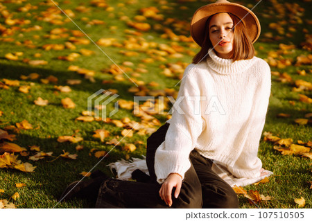 Autumn woman. Young cheerful woman in white sweater and hat walks in the nature park on a sunset. 107510505