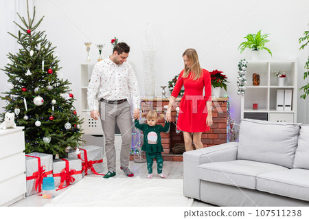 Mom and dad hold hands with their little daughter while standing by the Christmas tree. 107511238