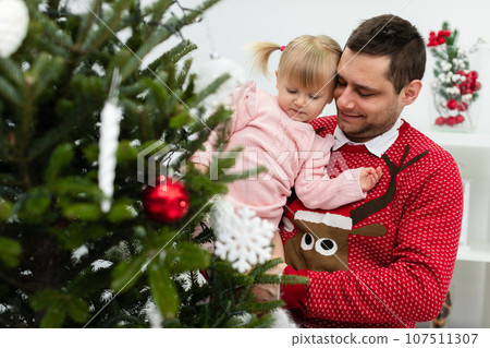 Dad and daughter decorate Christmas tree together by hanging ornaments on it Dad and daughter decorate Christmas tree together by hanging ornaments on it 107511307
