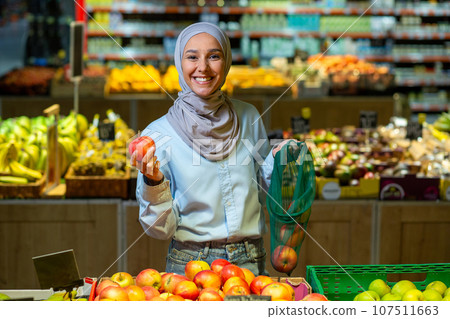 Portrait of a female buyer in a supermarket, a Muslim woman in a hijab is smiling happy and looking at the camera, choosing apples and fruits in a large grocery store. Portrait of a female buyer in a supermarket, a Muslim woman in a hijab is smiling happy and looking at the camera, choosing apples and fruits in a large grocery store. 107511663