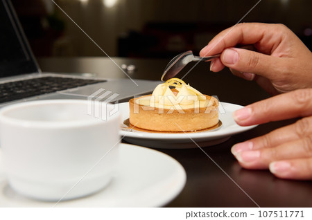 Close-up female hand holding a tea spoon over a French dessert with lemon custard, enjoying her coffee break while working on laptop. Cropped view. Copy advertising space Close-up female hand holding a tea spoon over a French dessert with lemon custard, enjoying her coffee break while working on laptop. Cropped view. Copy advertising space 107511771