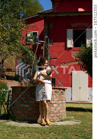 Beautiful happy mother holding her newborn baby standing by a well with water in the garden. Beautiful happy mother holding her newborn baby standing by a well with water in the garden. 107512355
