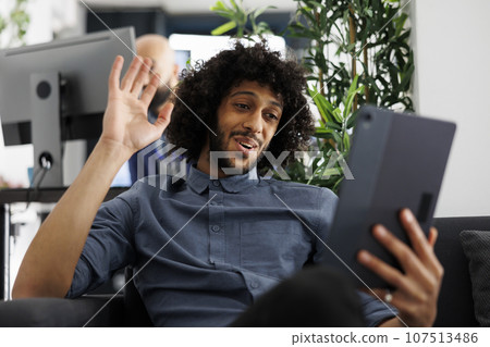 Start up company worker waving hi while talking in online meeting on digital tablet. Smiling arab project manager answering video conference call and greeting coworkers in office Start up company worker waving hi while talking in online meeting on digital tablet. Smiling arab project manager answering video conference call and greeting coworkers in office 107513486