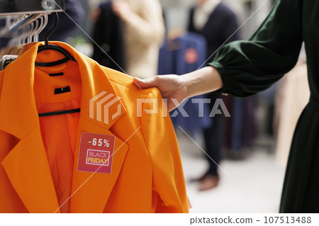 Female shopper choosing clothes in shopping center during Black Friday special offers buying clothing at discounted prices. Customer looking through rack with hanging apparel, hunting for bargains 107513488