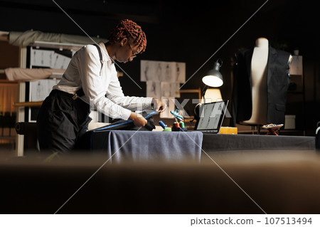 Hip level shot of suitmaker checking length size of unfinished custom made sartorial costume piece in atelier shop. Expert seamstress measuring exquisite suit garment for upcoming client comission 107513494