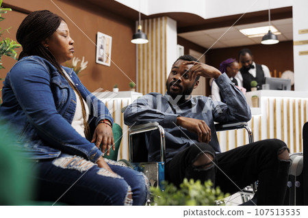 Tired African American guy in wheelchair arriving at luxury resort with wife, hotel guest with disability access needs waiting for check-in. Facilities for disabled tourists in hospitality industry 107513535