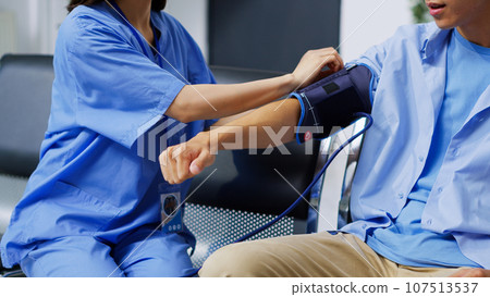 Asian nurse checking patient hypertension using medical tonometer during checkup visit appointment in hospital waiting area. Assistant examining elderly man blood pressure in reception 107513537