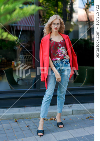 beautiful blonde woman in glasses dressed in red jacket and blue jeans posing on street in the city 107515722