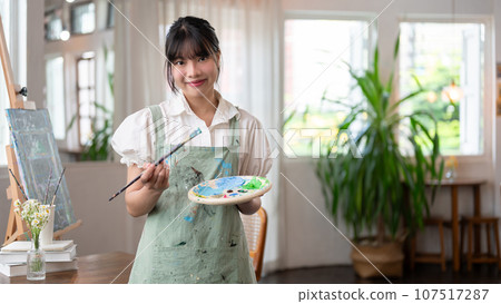 A charming Asian female artist holding a paint brush and a color palette, standing in her studio. 107517287