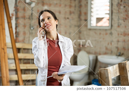 Positive female designer with papers talking on phone in building under renovation Positive female designer with papers talking on phone in building under renovation 107517506