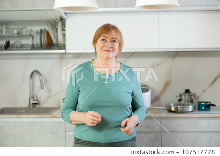 Smiling senior woman in casual clothes standing in home kitchen with open arms Smiling senior woman in casual clothes standing in home kitchen with open arms 107517778