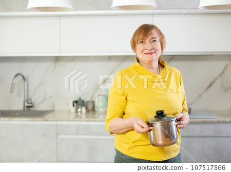 Elderly woman in casual clothes stands in front of table and holds stainless pan with lunch Elderly woman in casual clothes stands in front of table and holds stainless pan with lunch 107517866