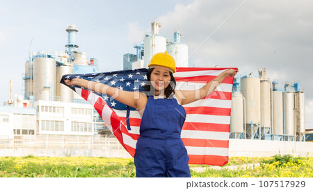 Portrait of positive girl in construction overalls with the USA flag on the background of modern factory 107517929