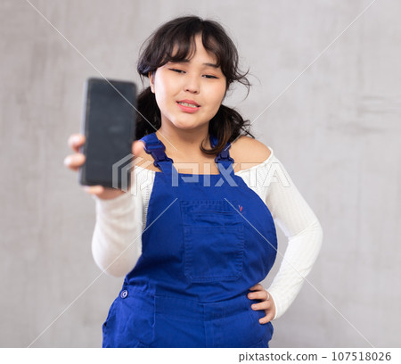 Positive girl in blue protective overalls showing modern smart phone with blank screen Positive girl in blue protective overalls showing modern smart phone with blank screen 107518026