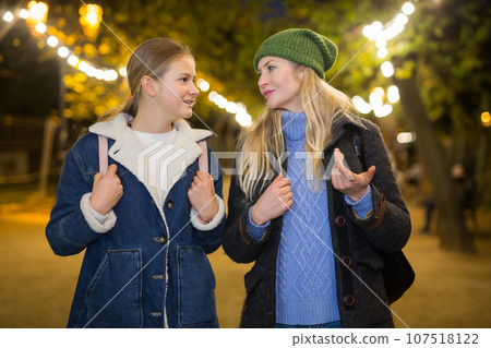 Positive woman walking with teenage daughter along evening city street Positive woman walking with teenage daughter along evening city street 107518122
