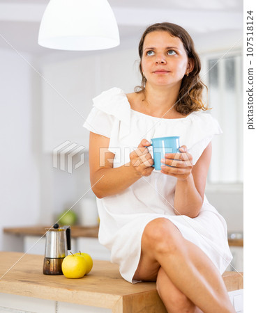 Young attractive girl posing in a white dress with a cup of coffee in modern kitchen Young attractive girl posing in a white dress with a cup of coffee in modern kitchen 107518124