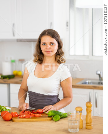 Positive woman is cutting vegetables for salad in kitchen at home Positive woman is cutting vegetables for salad in kitchen at home 107518127
