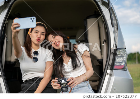 Two pretty Asian women are enjoying their road trip, using a phone to take selfies in an open trunk. 107522377