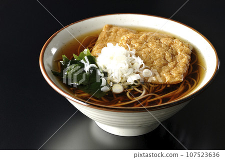 黑色背景下拍攝的Kitsune蕎麥麵,配上兩塊炸豆腐、海藻和大蔥。 黑色背景下拍攝的Kitsune蕎麥麵,配上兩塊炸豆腐、海藻和大蔥。 107523636