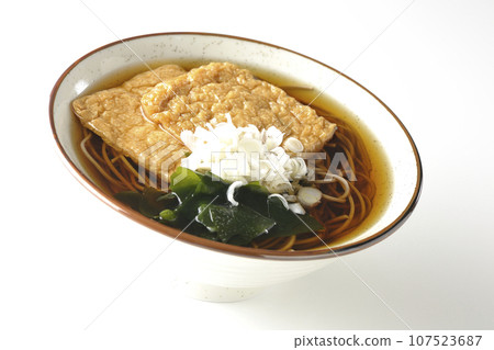 Kitsune soba noodles with two pieces of fried tofu, seaweed, and green onions are photographed against a white background. Kitsune soba noodles with two pieces of fried tofu, seaweed, and green onions are photographed against a white background. 107523687