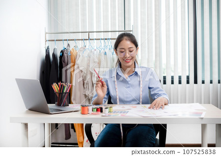 Beautiful young Asian fashion tailor with textile sewing accessories and entrepreneur designer sketches are full of bright colors on the desk with a laptop for creative ideas in the studio Beautiful young Asian fashion tailor with textile sewing accessories and entrepreneur designer sketches are full of bright colors on the desk with a laptop for creative ideas in the studio 107525838