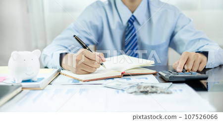 Man calculating her expenses on a notebook putting coin into piggy bank. Saving money with coins Step into a business that is growing to be successful and save for retirement concepts Man calculating her expenses on a notebook putting coin into piggy bank. Saving money with coins Step into a business that is growing to be successful and save for retirement concepts 107526066