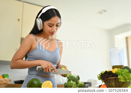 Beautiful woman in sportswear cutting broccoli while preparing healthy, vegetarian food in kitchen 107526564