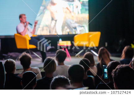 Crowd sitting in hall against blurred stage, while speaker showing presentation on big screen and speaking into mic during seminar 107527424