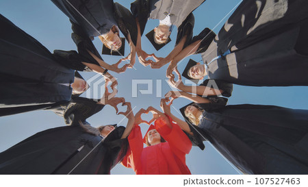 A group of seven college graduates make a heart shape from their hands while standing in a circle wearing robes. 107527463