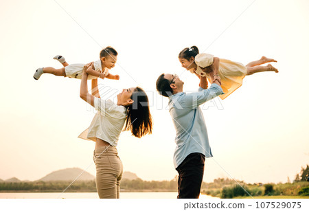 In a sunny park, a father and mother throw their happy son and daughter up in the air, while the children laugh and enjoy the playful freedom of flying. A moment of childhood happiness and family love 107529075