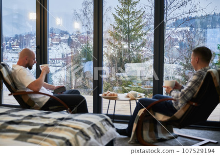 Focus on winter mountain landscape through panoramic windows in contemporary barn house. Two young men sitting on chair, holding cup of tea, enjoying view of magical scenery on blurred foreground. Focus on winter mountain landscape through panoramic windows in contemporary barn house. Two young men sitting on chair, holding cup of tea, enjoying view of magical scenery on blurred foreground. 107529194