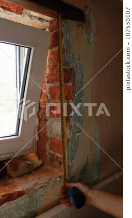 a vertical image where a builder, invisible in the frame, uses a tape measure to measure the height of a window opening in the process of installing a new small window in an old room a vertical image where a builder, invisible in the frame, uses a tape measure to measure the height of a window opening in the process of installing a new small window in an old room 107530107