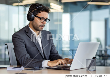 Serious thinking and concentrated man working with laptop inside office, man in headphones listening to audio books and podcasts at workplace. 107530146