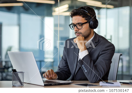Serious thinking and concentrated man working with laptop inside office, man in headphones listening to audio books and podcasts at workplace. 107530147