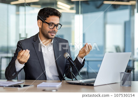 Young successful business coach recording podcast audio course about success and money, man using professional microphone and laptop while sitting at desk in middle of office. Young successful business coach recording podcast audio course about success and money, man using professional microphone and laptop while sitting at desk in middle of office. 107530154
