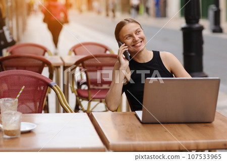 Successful smiling young business woman communication at smartphone during works on laptop at cafe outdoors. Concept of remote work from public place, digital freelance and modern lifestyle Successful smiling young business woman communication at smartphone during works on laptop at cafe outdoors. Concept of remote work from public place, digital freelance and modern lifestyle 107533965