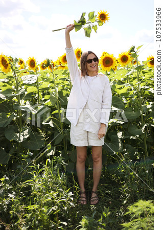 Smiling attractive young woman with short hair in sunglasses stands and relaxes at large bright sunflowers field. Stylish satisfied girl holds branch yellow sunflower looks at camera. Summer holiday. Smiling attractive young woman with short hair in sunglasses stands and relaxes at large bright sunflowers field. Stylish satisfied girl holds branch yellow sunflower looks at camera. Summer holiday. 107533966