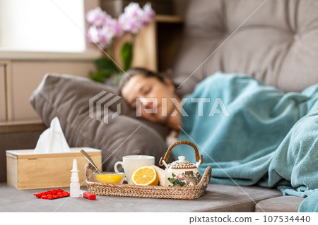 Flu epidemic. A sick woman sleeps under a blanket on the sofa. In front of her there is a tray with a teapot, lemon and honey. Nearby are pills, a thermometer, nasal spray and tissues. Blurry face. Flu epidemic. A sick woman sleeps under a blanket on the sofa. In front of her there is a tray with a teapot, lemon and honey. Nearby are pills, a thermometer, nasal spray and tissues. Blurry face. 107534440