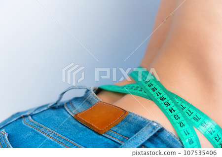 girl takes measurements of her body, on a gray background. Close-up of an unrecognizable slim young woman in jeans measuring her waist with a tape measure. Healthy lifestyle and weight loss concept 107535406