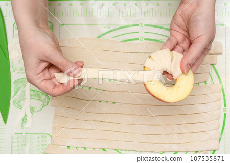 A young woman wraps a strip of puff pastry around an apple circle before baking it in the oven. A young woman wraps a strip of puff pastry around an apple circle before baking it in the oven. 107535871