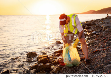 Earth Day. Volunteer man wearing vest in rubber gloves cleaning pebble wild beach. Copy space. Concept of ocean's pollution and save environment Earth Day. Volunteer man wearing vest in rubber gloves cleaning pebble wild beach. Copy space. Concept of ocean's pollution and save environment 107535929