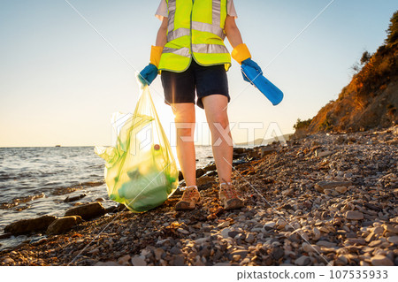 Low angle view at volunteer wearing vest and holds plastic garbage bag with bottle. Concept of environmental pollution and Earth Day 107535933