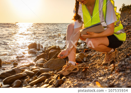 Close up of volunteer woman wearing vest and holds electronic tablet analyzing beach soil. In background is ocean, coast and sunset. Concept of inspection environmentalist and pollution Close up of volunteer woman wearing vest and holds electronic tablet analyzing beach soil. In background is ocean, coast and sunset. Concept of inspection environmentalist and pollution 107535934