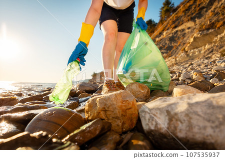 Bottom view of woman volunteer's hand in rubber gloves takes plastic bottle from pebble wild coast. Concept of ocean pollution 107535937