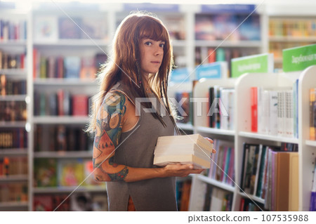Young thoughtful Caucasian woman with tattoo holds pile of books posing in bookstore. Concept of education and shopping in book shop Young thoughtful Caucasian woman with tattoo holds pile of books posing in bookstore. Concept of education and shopping in book shop 107535988