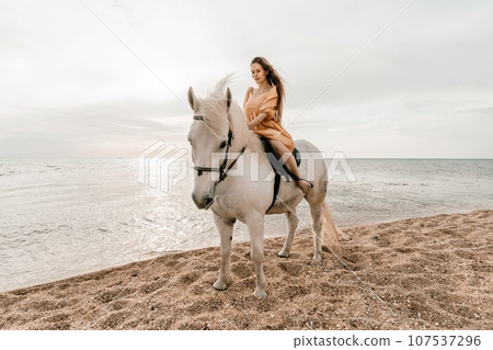 A woman in a dress stands next to a white horse on a beach, with the blue sky and sea in the background. A woman in a dress stands next to a white horse on a beach, with the blue sky and sea in the background. 107537296