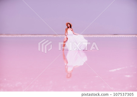 Woman in pink salt lake. She in a white dress and hat enjoys the scenic view of a pink salt lake as she walks along the white, salty shore, creating a lasting memory. Woman in pink salt lake. She in a white dress and hat enjoys the scenic view of a pink salt lake as she walks along the white, salty shore, creating a lasting memory. 107537305